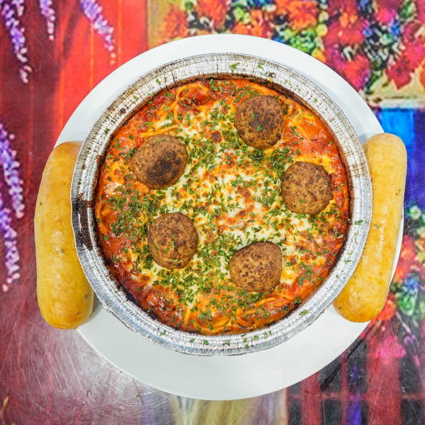 Baked spaghetti with meatballs and melted cheese in a foil pan, served with two breadsticks.
